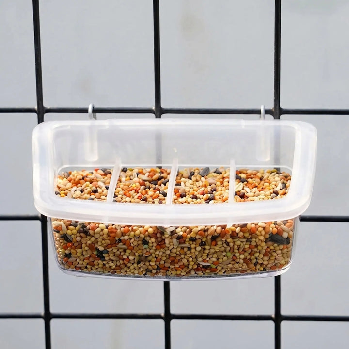Four-section clear feeder full of mixed seed, hung by two metal hooks on black cage wire. 3