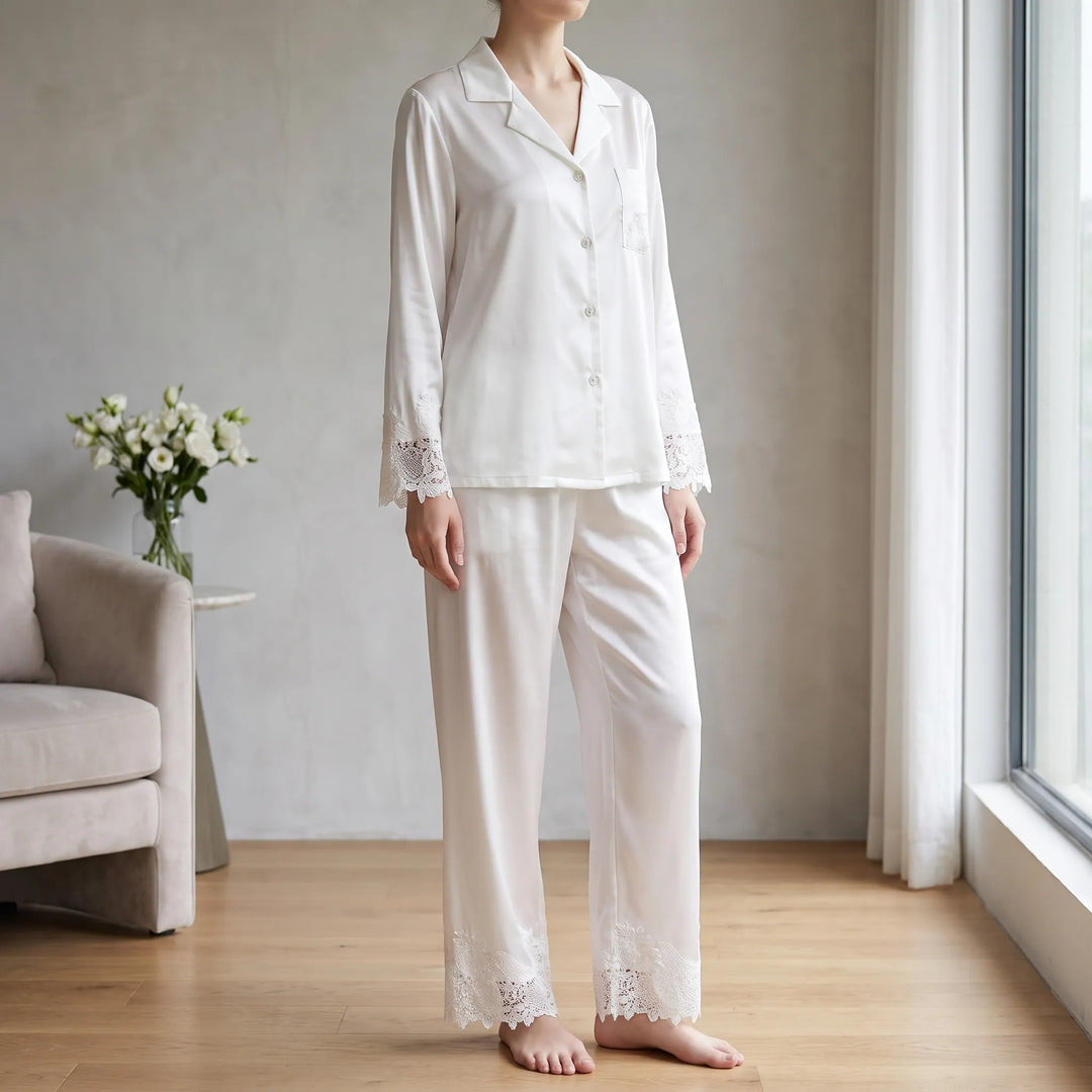 Model in white satin pajama set with lace cuffs and hems standing barefoot in a bright living room by a window. 1