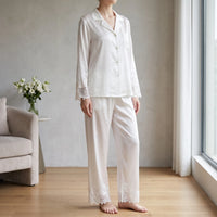Model in white satin pajama set with lace cuffs and hems standing barefoot in a bright living room by a window. 1