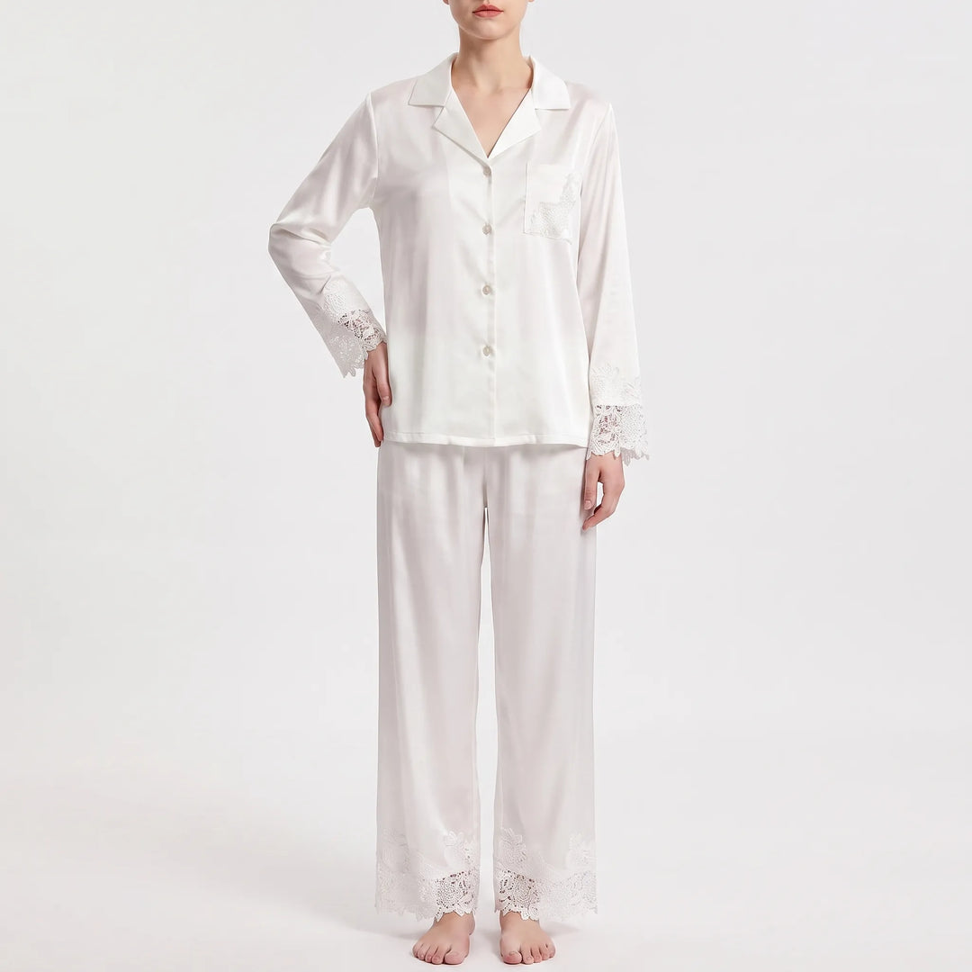 Front view on a plain background of the white satin pajamas with lace-trimmed sleeves, pocket and trouser hems, one hand on hip. 2