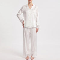 Front view on a plain background of the white satin pajamas with lace-trimmed sleeves, pocket and trouser hems, one hand on hip. 2