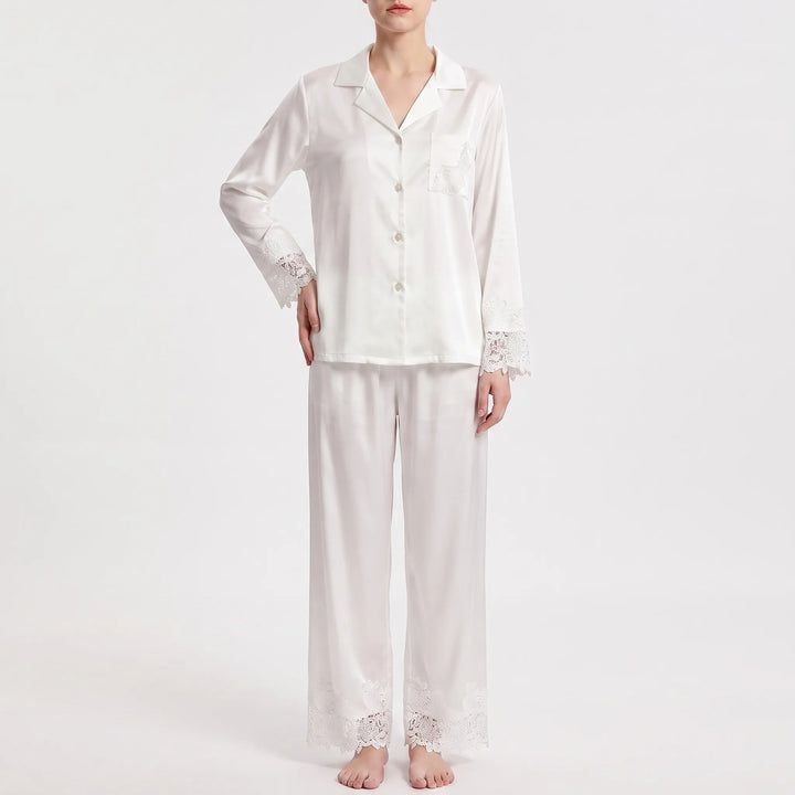 Front view on a plain background of the white satin pajamas with lace-trimmed sleeves, pocket and trouser hems, one hand on hip. 2