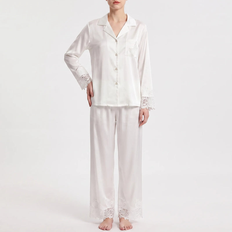 Front view on a plain background of the white satin pajamas with lace-trimmed sleeves, pocket and trouser hems, one hand on hip. 2