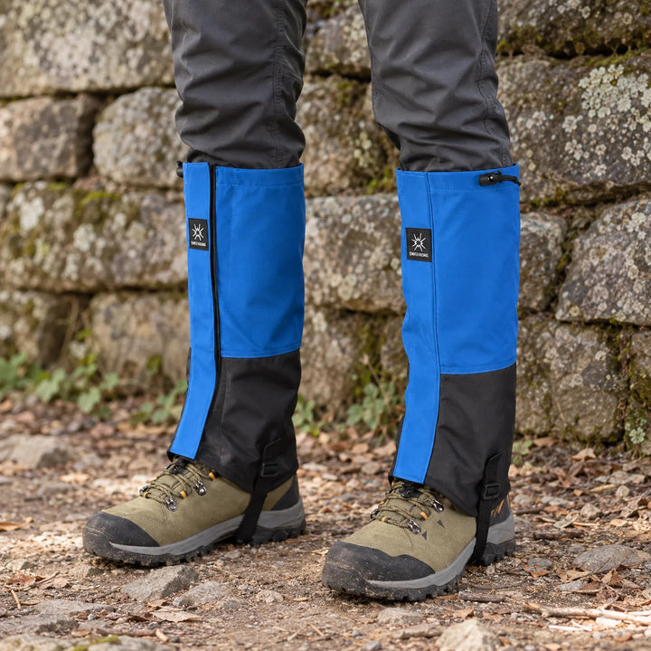 Blue and black gaiters with side zips worn over olive hiking boots on a leaf-strewn trail beside a lichen-covered stone wall. 1