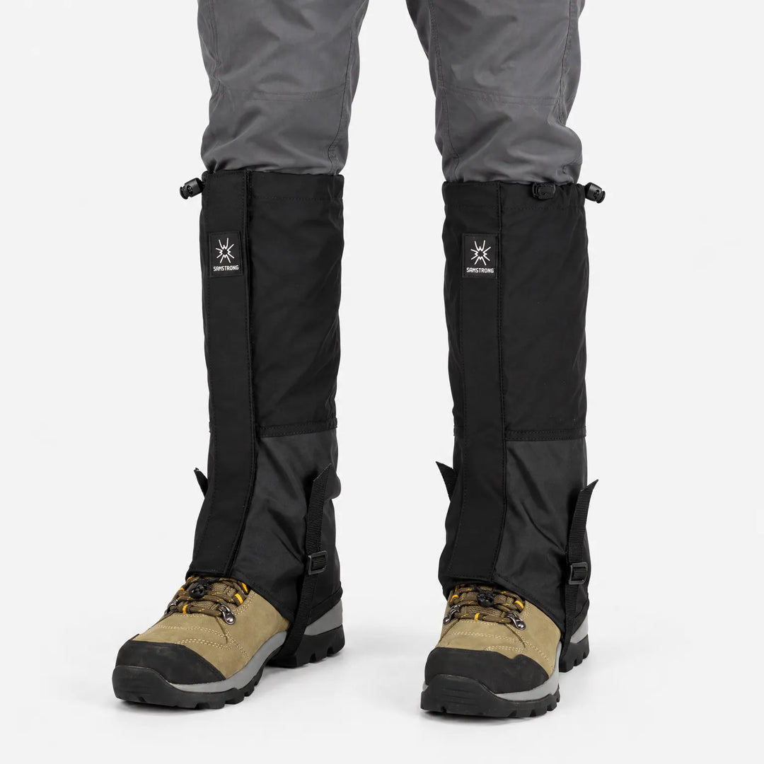 All-black gaiters with drawcord tops and ankle straps, worn over grey trousers and tan hiking boots, shown from the front. 8