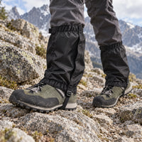 Black ripstop gaiters worn over hiking boots and trousers on a rocky mountain trail with snowy peaks behind. 1