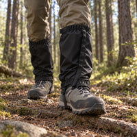 Black gaiters worn over muddy hiking boots on a forest trail, seen from a low angle among pine trees. 3