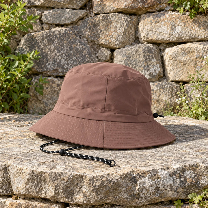Brown bucket hat resting on a sunlit stone ledge, side view showing the stitched brim and drawcord with toggle. 3