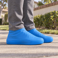 Person in grey jeans wearing blue stretchy rubber shoe covers outdoors on a sunlit pavement, seen from a low side angle. 1