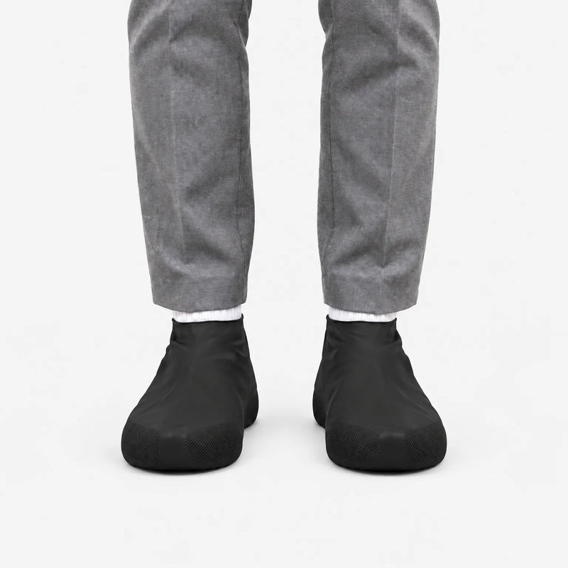 Front view of black rubber shoe covers worn with grey trousers and white socks, showing the textured grip at the toes. 4