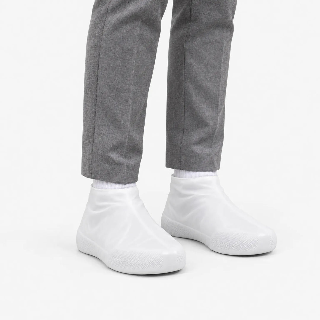 Angled view of translucent white rubber shoe covers worn with grey trousers, showing the textured toe area and snug ankle fit. 5