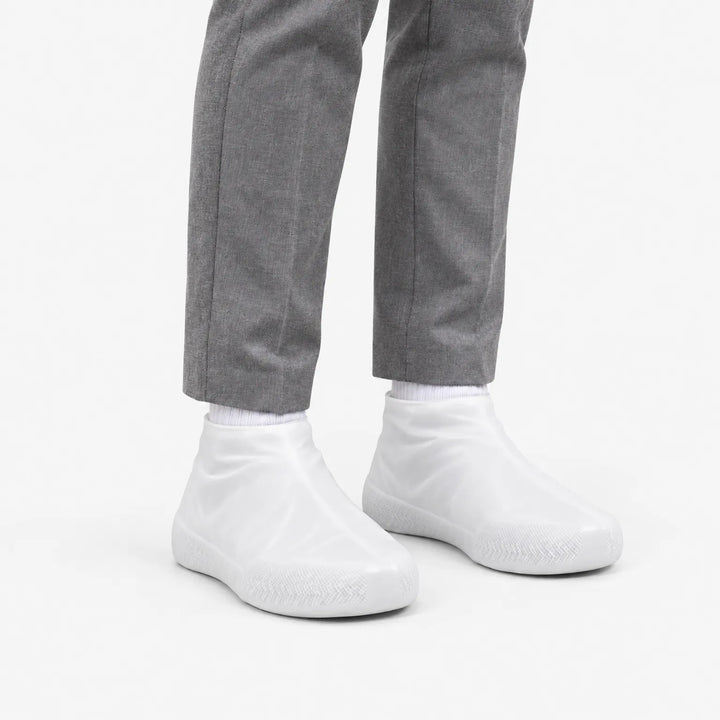 Angled view of translucent white rubber shoe covers worn with grey trousers, showing the textured toe area and snug ankle fit. 5