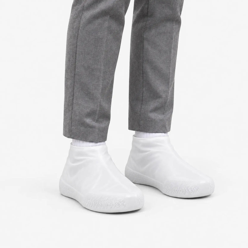 Angled view of translucent white rubber shoe covers worn with grey trousers, showing the textured toe area and snug ankle fit. 5