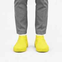Front view of bright yellow rubber shoe covers worn with grey trousers and white socks against a plain white background. 7