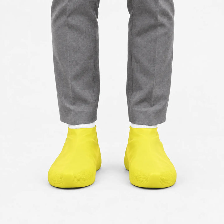 Front view of bright yellow rubber shoe covers worn with grey trousers and white socks against a plain white background. 7