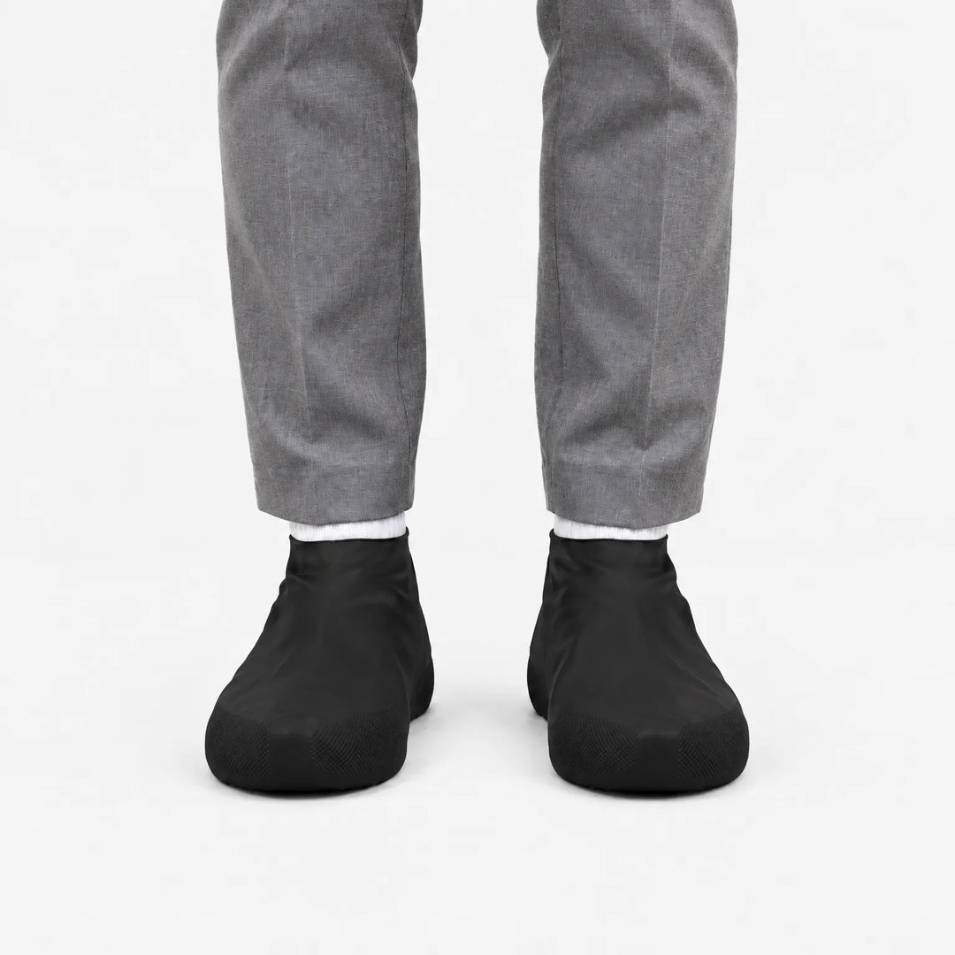 Another front view of black rubber shoe covers worn with grey trousers and white socks, with textured toe grip visible. 9