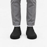 Another front view of black rubber shoe covers worn with grey trousers and white socks, with textured toe grip visible. 9