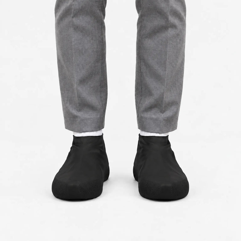 Another front view of black rubber shoe covers worn with grey trousers and white socks, with textured toe grip visible. 9