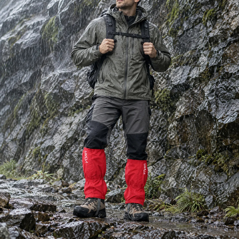 Hiker in a rain jacket and backpack wears red gaiters over hiking boots while standing on a wet rocky trail beside a rock face. 1