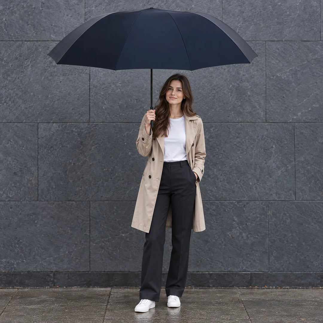 Woman in a beige trench coat and white sneakers holding a large navy umbrella against a grey stone wall. 1
