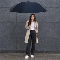 Woman in a beige trench coat and white sneakers holding a large navy umbrella against a grey stone wall. 1