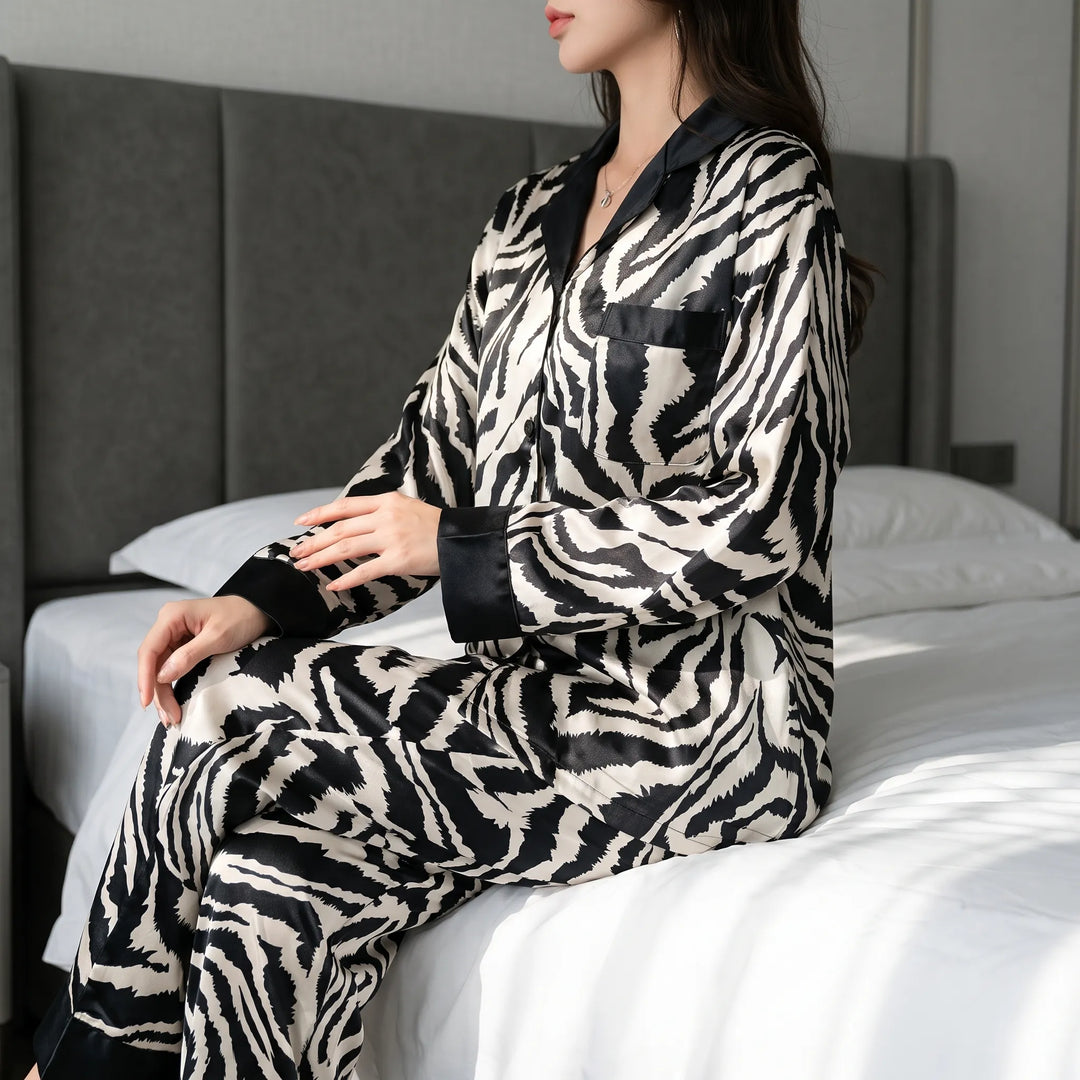 Woman sitting on the edge of a bed in the long-sleeved zebra-print satin pyjama set with black collar, cuffs and chest pocket. 1