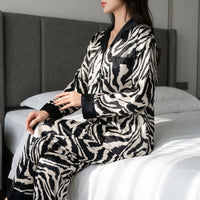 Woman sitting on the edge of a bed in the long-sleeved zebra-print satin pyjama set with black collar, cuffs and chest pocket. 1