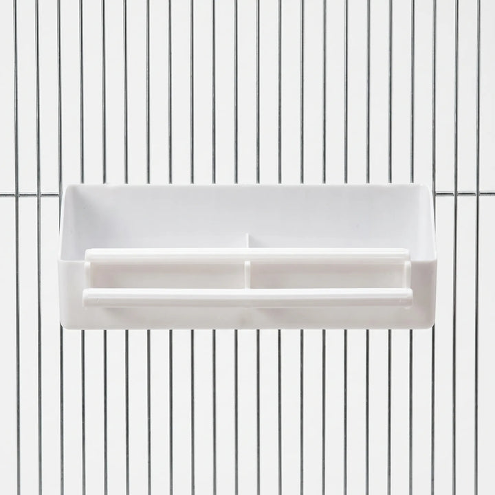 Front view of a white plastic two-compartment feeder with two perches, hung on the vertical bars of a metal cage. 1