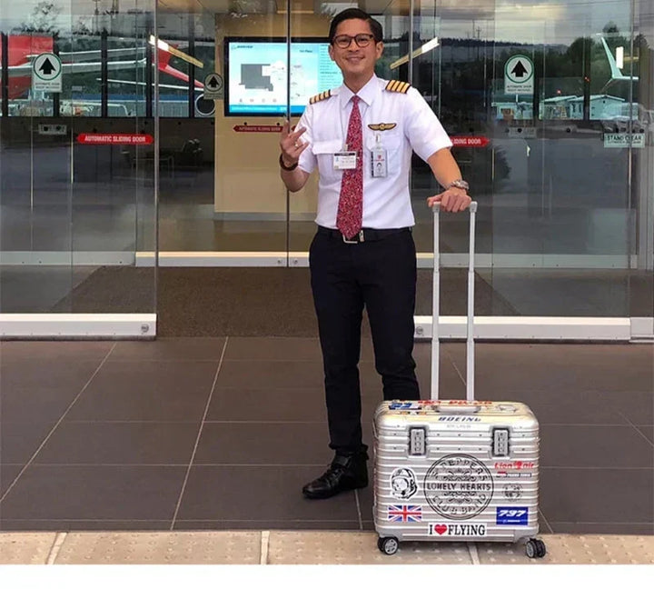 Smiling airline pilot in uniform stands beside a silver case covered in travel stickers in an airport entrance 4