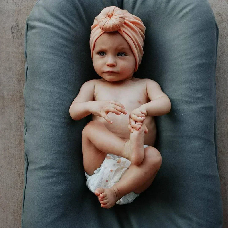 Baby in a peach knotted hat holding one foot, lying in a grey-blue lounger 3
