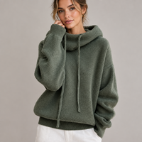 Smiling model in a sage green knit jumper with drawstring roll neck, hand at cheek, white trousers 1