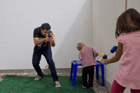 Man, girl and toddler play outdoors on a small lawn, two aiming blue and yellow blasters 10