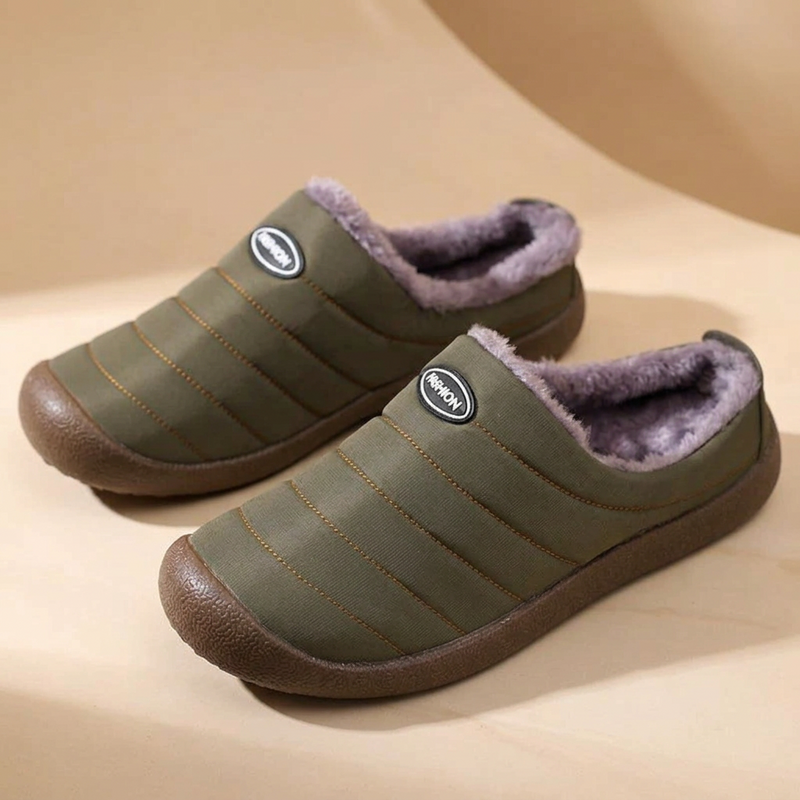 Pair of olive green quilted slippers with purple-grey fleece lining on a beige curved backdrop 17