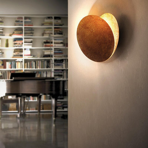 Side view of the round lamp with its rear disc angled open, beside a bookshelf and piano 4
