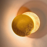 Gold-finish wall lamp with offset discs, the rear disc lit, on a textured pale wall 12