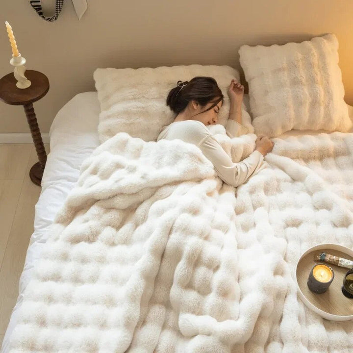 Woman sleeping under a cream bubble-textured plush blanket, with matching cushions and a lit candle on a tray 1