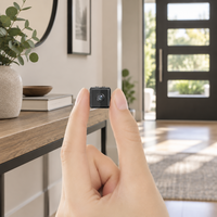Tiny black cube camera held between finger and thumb in a bright hallway beside a console table 1