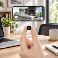 Tiny square black camera held between finger and thumb above a desk, with a phone showing a live living room view 1